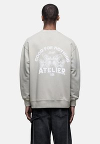 Lichtgrijze sweatshirt met een oversized pasvorm, ribbels aan de mouwen en zoom. Voorzien van een vlinderafbeelding en de tekst "GOOD FOR NOTHING ATELIER 1986" op de achterkant.