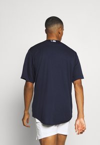 Laivastonsininen lyhythihainen T-paita, jossa on pyöreä pääntie, valkoinen logo takana ja rento istuvuus. Sileä, hengittävä kangaspinta.