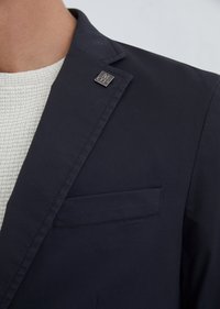 Navy blazer with a tailored fit, featuring a sleek lapel, single pocket, and a metal logo pin on the collar. Textured light gray sweater underneath.