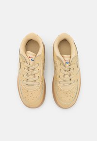 Beige lädersneakers med perforerad design, med grå detaljer, bruna skosnören och en sula av gummirubber. Nike-logotyp i blått och rött.