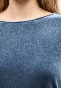 Κοντινό πλάνο ενός ατόμου που φοράει απαλό μπλε βελούδινο πάνω μέρος με στρογγυλή λαιμόκοψη, που δείχνει τον λαιμό και την περιοχή του άνω στήθους.