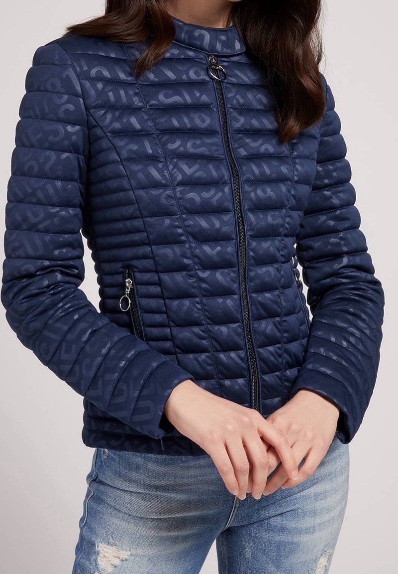 Femme portant une veste matelassée ajustée bleu marine avec fermeture éclair et lettres à motif subtil, associée à un jean bleu clair déchiré, les mains légèrement jointes.