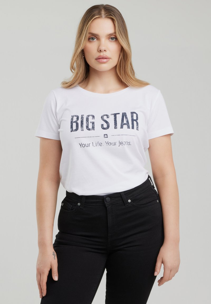 Žena se světlými vlasy, která má na sobě bílé tričko s nápisem „BIG STAR Your Life. Your Jeans.“ a černé džíny s vysokým pasem, stojící před jednobarevným pozadím.
