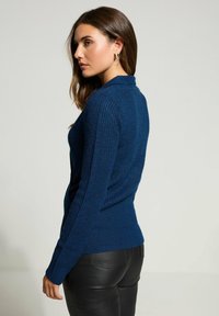 Donna con lunghi capelli castani che indossa un maglione blu a trama e pantaloni in pelle nera, in piedi di lato contro uno sfondo neutro.