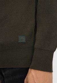 Dunkelolivgrüner gerippter Pullover mit strukturiertem Stoff, ausgestattet mit einem quadratischen Logo-Patch am linken Ärmel und einem taillierten Bündchendesign.