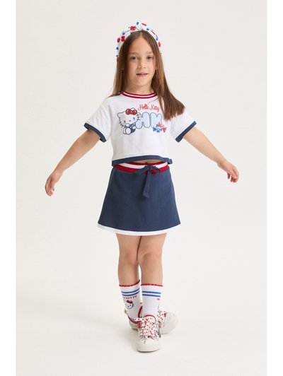 Mlada deklica v majici Hello Kitty, mornarsko modri krilu z rdečim in belim robom, ujemajoče nogavice, cvetlične čevlje in pisano trak za lase, stoji z razprtimi rokami.