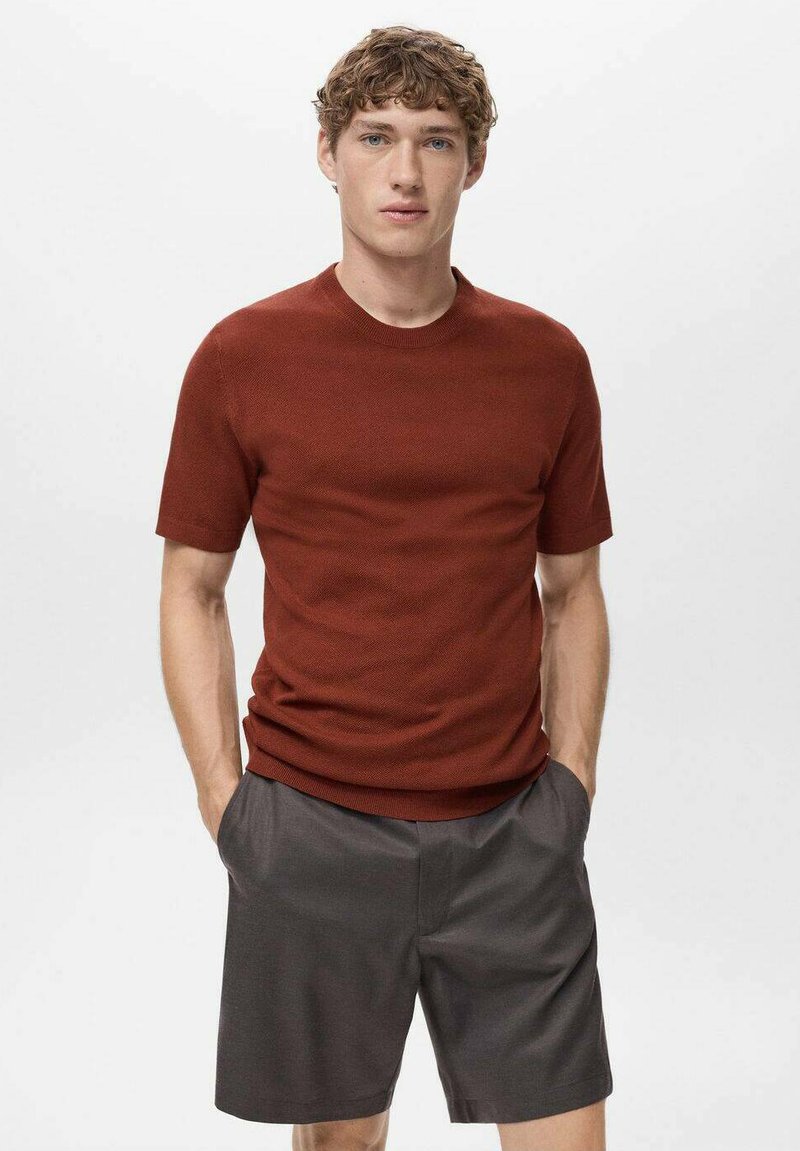 Tricou scurt de culoare ruginie, realizat dintr-o țesătură texturată, asortat cu pantaloni scurți gri închis. Modelul stă cu mâinile în buzunare, pe un fundal simplu.