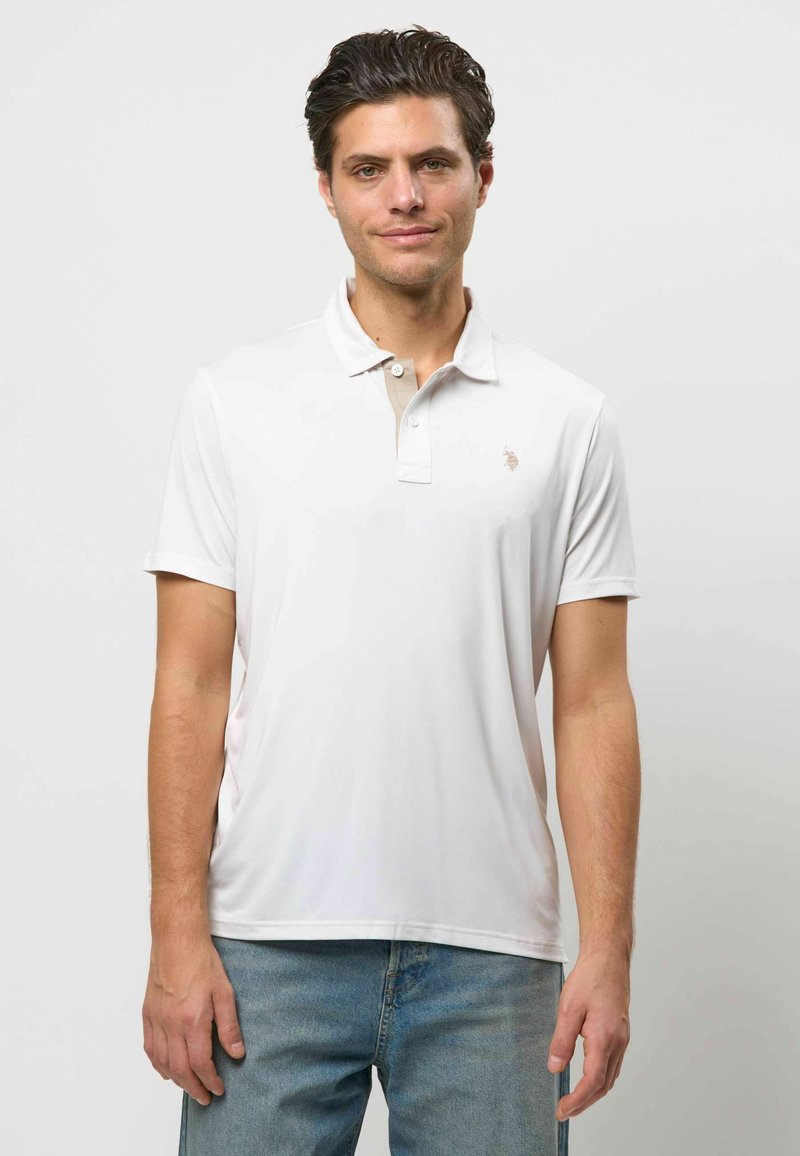 Homme aux cheveux courts et foncés, portant un polo blanc et un jean bleu, debout devant un fond clair uni.