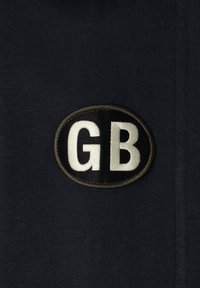 Oválny textilný nášivka s bielymi písmenami "GB" na čiernom pozadí, prišitá na tmavú textúrovanú tkaninu.