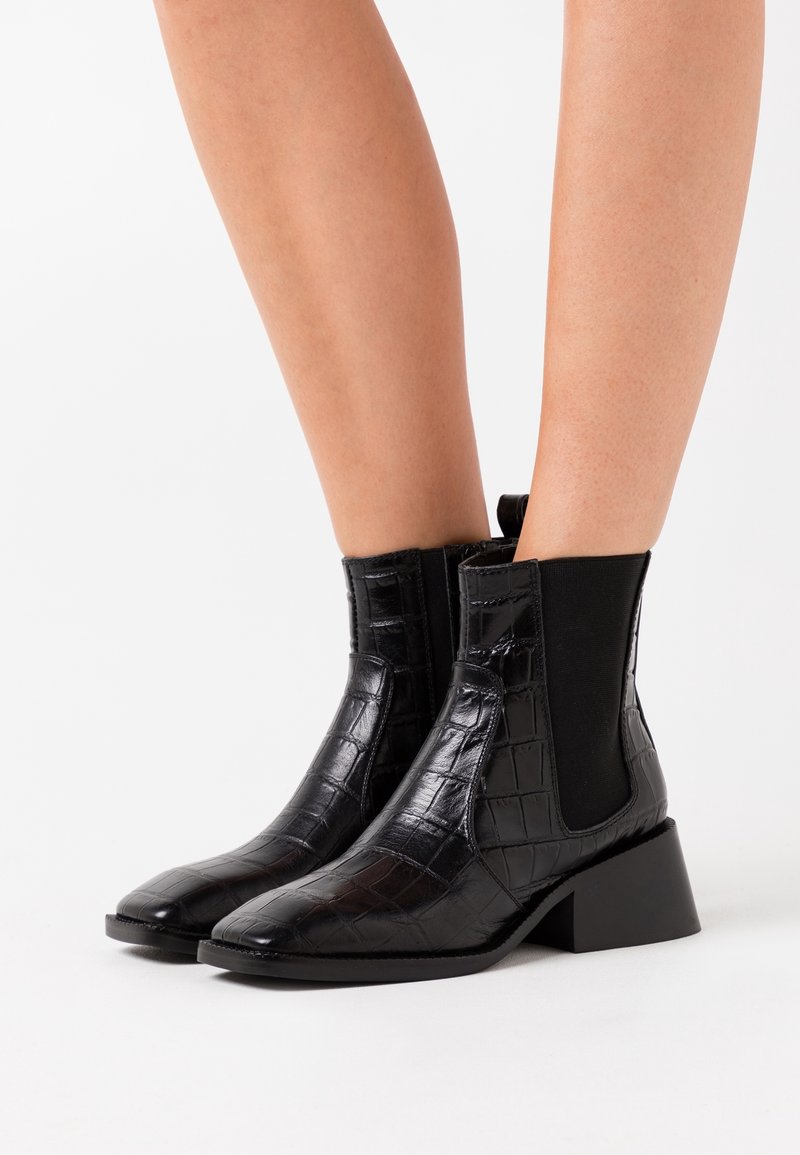 Bottes en cuir noires mi-mollet avec motif crocodile, panneaux latéraux élastiques et talons blocs, portées sur des jambes nues sur fond blanc.