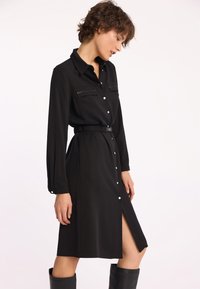 Czarna sukienka koszulowa z długimi rękawami, zapięciem na guziki i dwiema kieszeniami na piersi. Posiada paski w talii oraz wysoki rozcięcie z boku. Gładka faktura materiału.