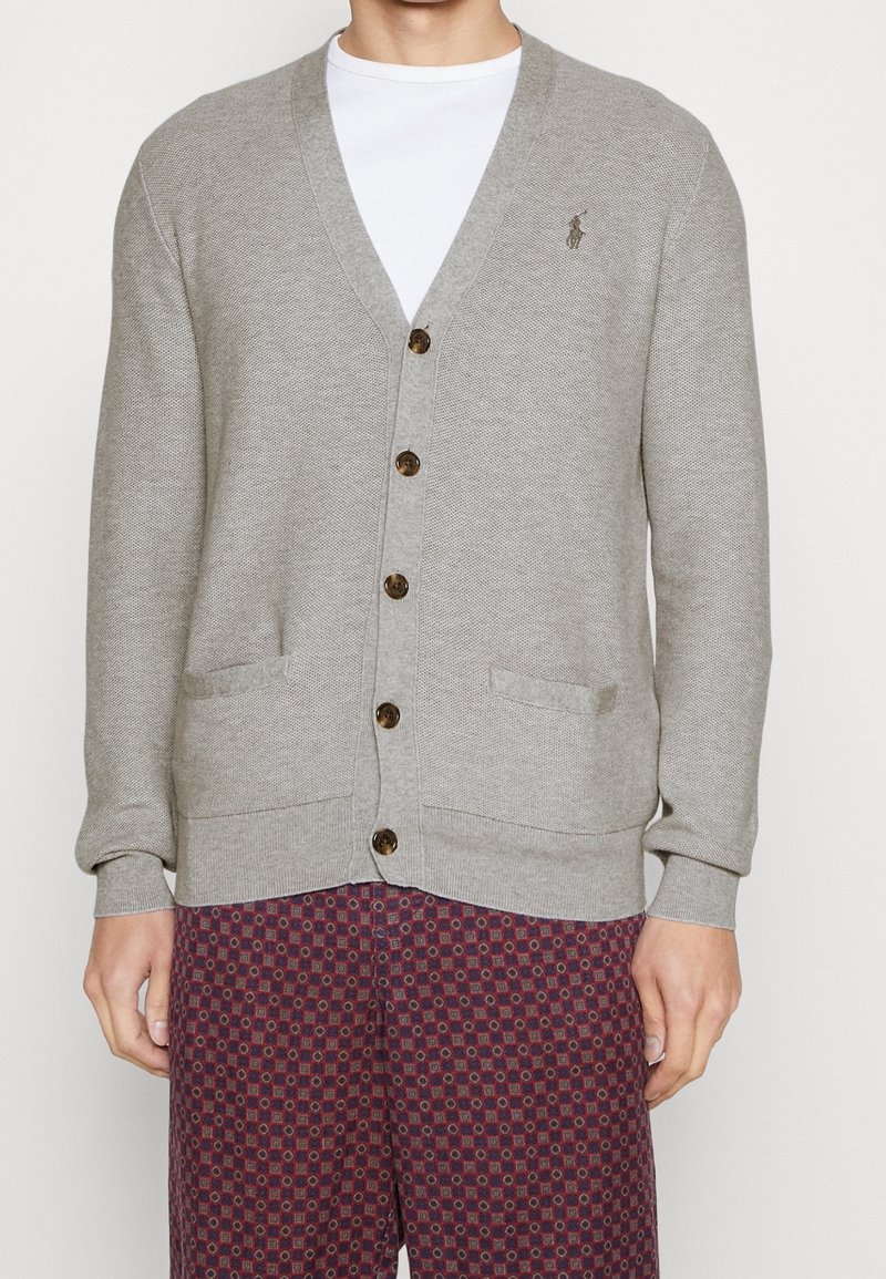 Mann trägt hellgrauen, geknöpften Cardigan über weißem Hemd und rot gemusterte Hose, steht vor einem schlichten weißen Hintergrund.