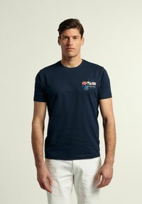 Camiseta de algodón azul marino con un gráfico de navegación y texto, de manga corta y cuello redondo. Se lleva con pantalones blancos.