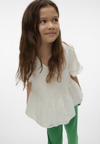 Top in cotone bianco con maniche corte, motivi ad occhio di pernice, scollatura a V e vita a peplo. Abbinato a pantaloni verdi, su uno sfondo neutro.