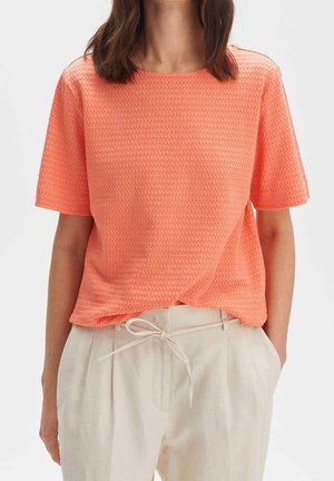 Frau trägt ein strukturiertes kurzärmliges Korallenoberteil, das in eine hoch taillierte, cremefarbene Hose mit gebundenem Gürtel gesteckt ist.