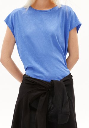 Kobieta nosząca niebieski t-shirt z krótkim rękawem i czarną bluzę zawiązaną wokół talii na białym tle.