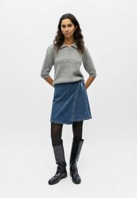 Femme aux cheveux bouclés portant un pull en maille gris, une jupe portefeuille en denim, des collants noirs et des bottes en cuir noires montantes, sur un fond uni.