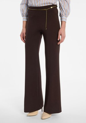 Frau trägt dunkelbraune High-Waist-Hose mit weitem Bein, dünnen goldenen Kettengürtel, gestreifte Bluse und beige spitze Absatzschuhe, steht vor weißem Hintergrund.
