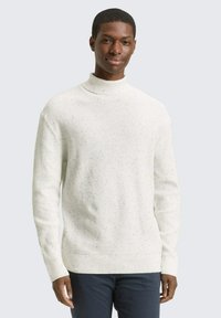 Pull à col roulé blanc à côte, présentant un design texturé avec des éclats. Des manches longues et une coupe décontractée complètent le look.
