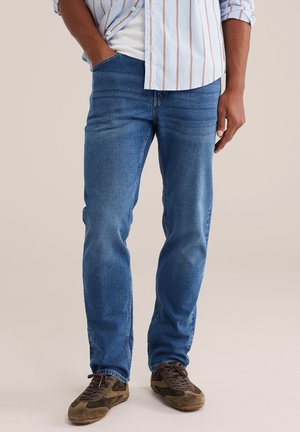 Homme portant un jean bleu, des baskets marron et vertes, une main dans la poche et une chemise rayée sur un t-shirt blanc.