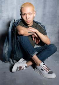 Sneakers bianchi con lacci marroni e dettagli grigi, indossati con jeans blu arrotolati e una giacca di jeans, su uno sfondo grigio.