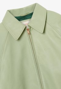 Hellgrüne Lederjacke mit spitzem Kragen und goldenem Reißverschluss, mit teilweiser Markenprägung auf der Brust.
