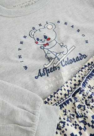 Grauer Sweatshirt mit einem cartoonhaften Bären auf Skiern und dem Text "Winter Ski Saison Aspen, Colorado". Kombiniert mit weißen Shorts mit blauen geometrischen Mustern.