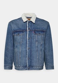 Blå denimjacka med vit sherpa-krage, knäppning fram, två bröstfickor med lock och sidfickor, lagt på en vit bakgrund.