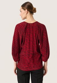 Rød mønstret bluse med et tekstureret rutedesign, batwing-ærmer, rund halsudskæring og en nøglehulslukning bagpå.