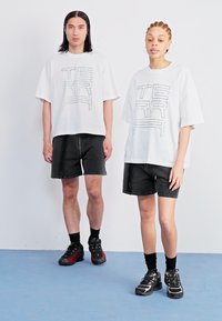Baltos per didelio dydžio t-shirts su juodais grafikos tekstais "TERR" didelėmis raidėmis. Juodi džinsiniai šortai ir juodi sportiniai bateliai užbaigia aprangą.