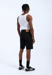 Pantalones cortos de mezclilla negra con corte recto, combinados con una camiseta sin mangas blanca, calcetines negros y zapatillas negras con rayas blancas. Se muestra la vista trasera.