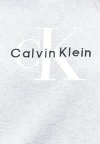 Tissu gris clair avec de grandes lettres blanches "CK" et un texte plus petit noir "Calvin Klein" brodé par-dessus les lettres.