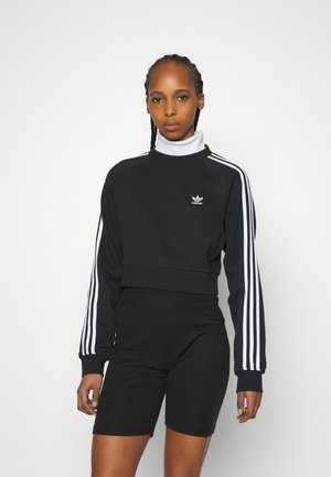 Felpa nera cropped con dettagli bianchi a tre strisce sulle maniche e un piccolo logo marchiato. Abbinata a pantaloni corti neri, design minimale.