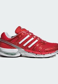 Chaussure de sport rouge avec des bandes blanches, dessus en maille, semelle rembourrée et coussin d’air visible au talon, vue de profil sur fond blanc.