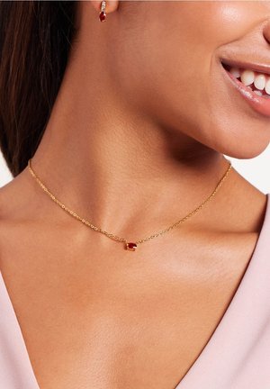 Gouden ketting met een kleine rode edelsteen hanger. Soepele textuur, lichtgewicht ontwerp, gedragen op een blote nek tegen een lichte top.