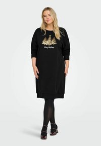 Žena u crnoj haljini dugih rukava s zlatnim božićnim drvcima i natpisom "Sretan Božić", crnim tajicama i tamnocrvenim Mary Jane cipelama, stoji.