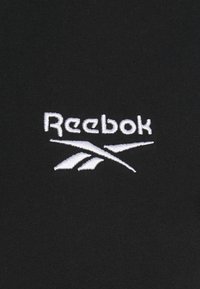 Černá látka s bílou vyšívanou logem Reebok, s elegantními písmeny a ostrými akcenty, vytvářející kontrastní designový prvek.