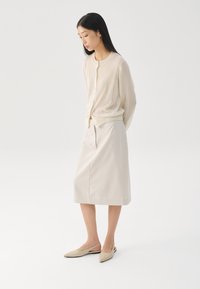 Cárdigan de punto beige sobre una falda de crema clara, con zapatos de punta tipo slip-on. Texturas suaves y un diseño sencillo, con un escote redondeado y botones.