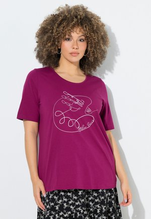 Kvinde med krøllet hår har øreringe i hoops, en lilla skjorte med hvidt abstrakt hånddesign og et sort skørt med hvide skriftmønstre.