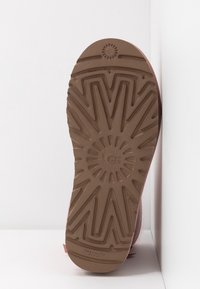 Semelle en caoutchouc marron avec un motif en zigzag texturé, comportant un logo circulaire au centre. La partie supérieure rose est légèrement visible.