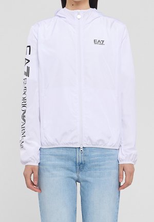 Giacca con cappuccio bianca con cerniera e logo EA7 Emporio Armani sul petto e sulla manica, indossata con jeans azzurri, mostrata dal collo ai fianchi.