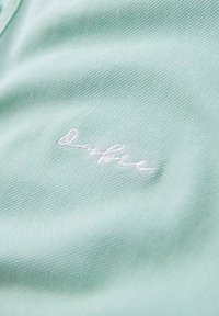 Tessuto verde menta chiaro con una trama testurizzata, caratterizzato da un logo in bianco ricamato sul lato inferiore. Vista ravvicinata del materiale.