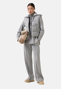 Kobieta ubrana w jasnoszary pikowany bezrękawnik na dopasowanej koszuli i szerokich spodniach, trzyma beżową kurtkę z owczej wełny, stoi na tle białego tła.