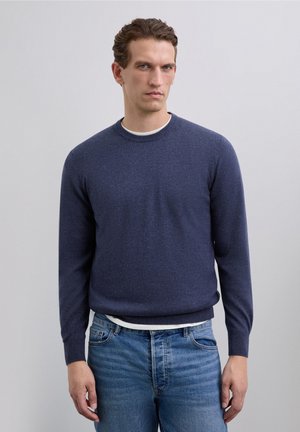 Hombre con cabello corto y ondulado, vistiendo un suéter de cuello redondo azul marino sobre una camiseta blanca y jeans azules, de pie frente a un fondo claro y sencillo.