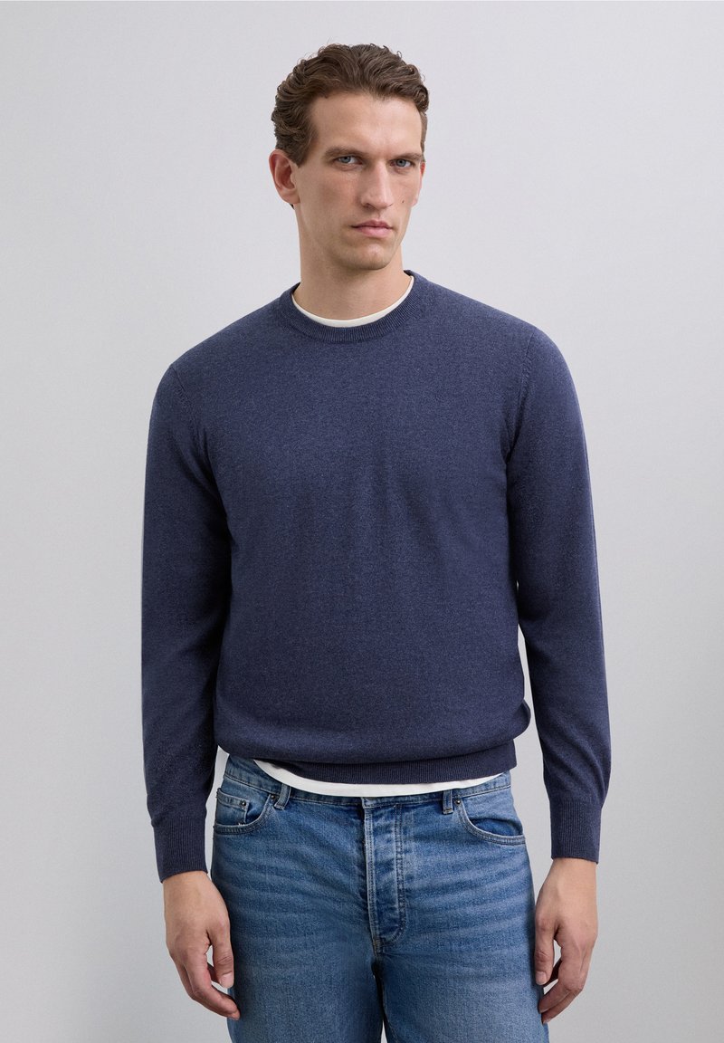 Mand med kort bølget hår iført en navy crewneck sweater over en hvid skjorte og blå jeans, stående foran en plain lys baggrund.