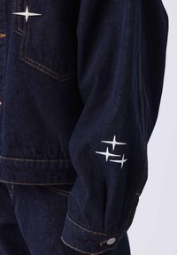 Giacca in denim scuro con un design strutturato, caratterizzata da ricami bianchi a forma di stella sulle maniche e cuciture gialle a contrasto.