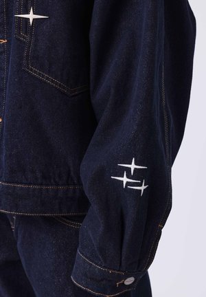Giacca in denim scuro con un design strutturato, caratterizzata da ricami bianchi a forma di stella sulle maniche e cuciture gialle a contrasto.