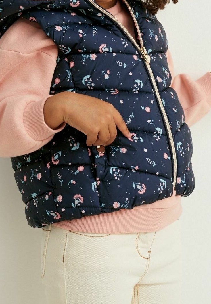 Enfant portant un gilet matelassé à fleurs bleu marine avec fermeture éclair et un sweat-shirt rose, mettant la main dans la poche du gilet.