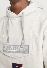Sudadera beige hecha de tela suave, con un cordón gris, detalles de parche con el texto "Napapijri" y un emblema de bandera.