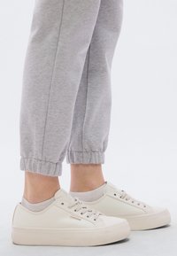 Balti sporta apavi ar gludu, teksturētu virsmu, apaļu purngalu un platu zoli, saskaņoti ar gaismi pelēkām, elastīgām potīšu sporta biksēm.